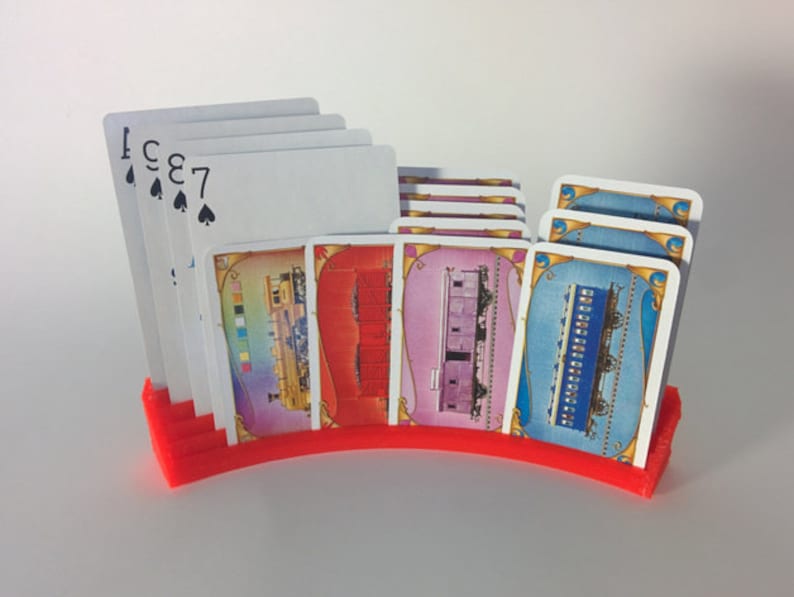 May include: A red plastic card holder with a curved design holds a deck of playing cards and several train-themed cards. The cards feature images of trains and numbers.