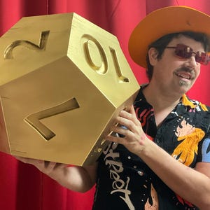 May include: A large, gold-coloured, twenty-sided die with carved numerals. The die is held in front of a red curtain. The numbers are carved into the surface of the die. The die is a prop for a game.