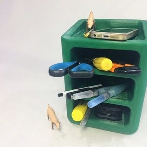 May include: A green, three-tiered organizer with various items stored inside. A smartphone, scissors, pens, and other small objects are visible. A small figurine of an animal is on the side. The organizer is made of a solid material.