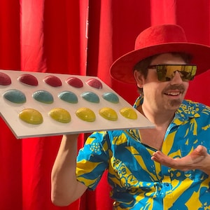 May include: A person holding a white board with colorful, round objects. The objects are arranged in rows of red, blue, and yellow. The person is wearing a red hat, sunglasses, and a blue and yellow patterned shirt.