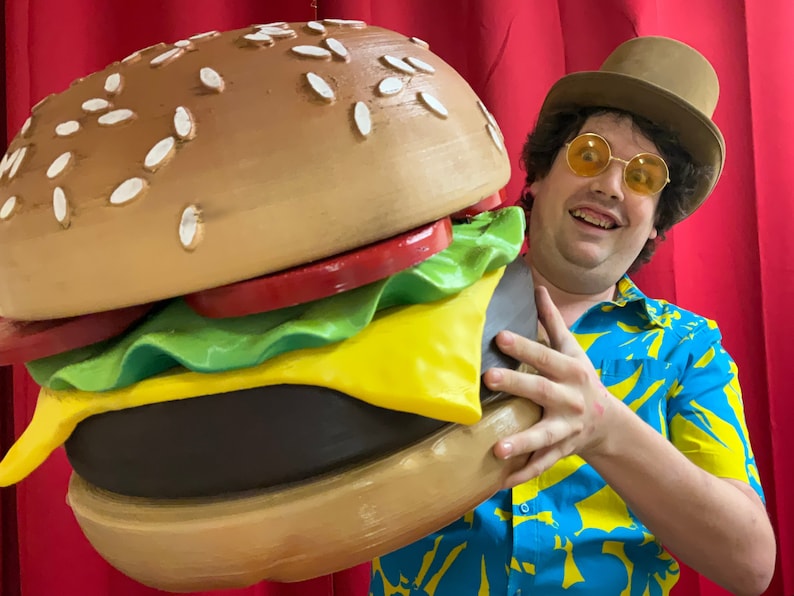 Puede incluir: Una persona que lleva una camisa hawaiana azul y amarilla y un sombrero de copa marr&oacute;n sostiene una gran hamburguesa de gran tama&ntilde;o. La hamburguesa tiene semillas de s&eacute;samo en el pan superior, lechuga, tomate, queso y una hamburguesa de carne.
