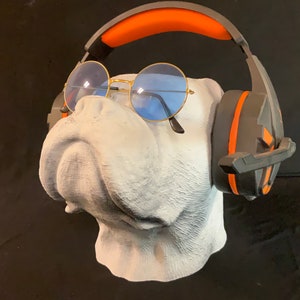 Può includere: Un busto di bulldog bianco con occhiali rotondi blu e cuffie nere e arancioni.