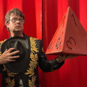 May include: A person holding a large, red, pyramid-shaped die with white numbers. The person is wearing a black jacket with gold embroidery and a black hat. The background is a red curtain.