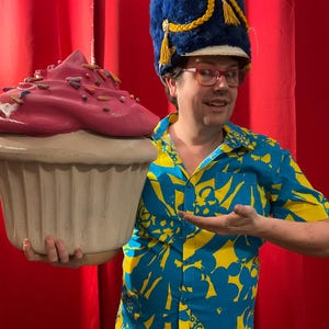 May include: A person holding a large cupcake prop with pink frosting and sprinkles. The person is wearing a blue and yellow patterned shirt, red glasses, and a blue hat with gold trim. The background is a red curtain.