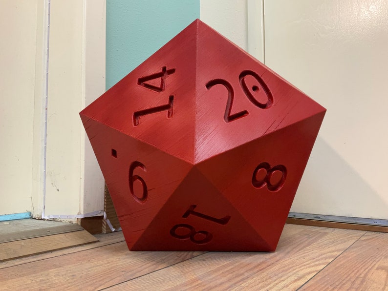 Puede incluir: Un dado de madera de veinte caras grande, rojo, con los n&uacute;meros 1, 4, 6, 8, 17 y 20 visibles.