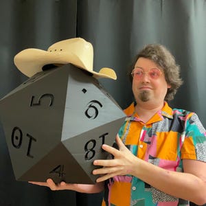 Pu&ograve; includere: Un grande dado nero a venti facce con numeri bianchi viene tenuto in alto. Un cappello da cowboy marrone chiaro &egrave; posto sopra il dado. La persona che tiene il dado indossa una camicia a maniche corte colorata con un motivo geometrico.