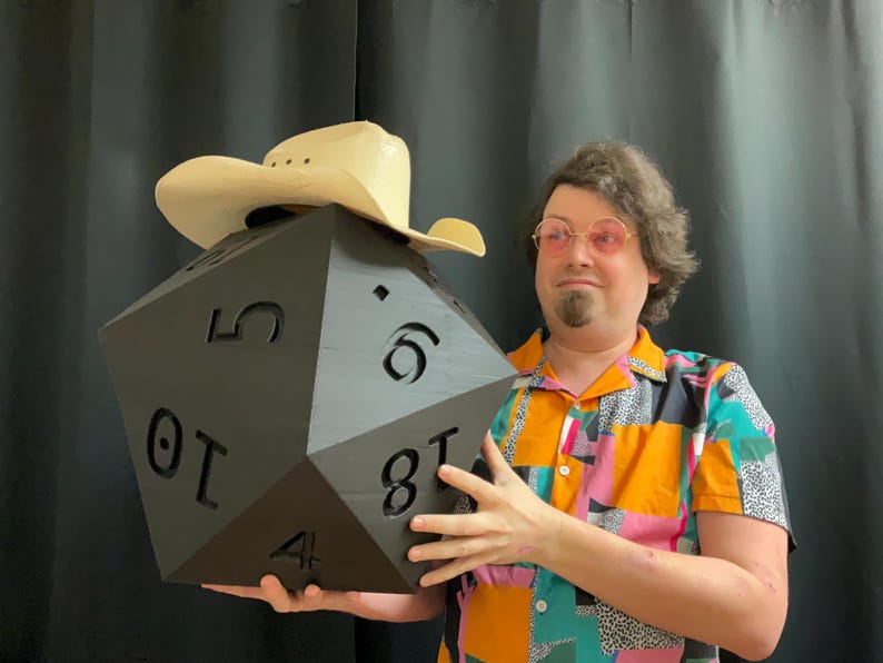Pu&ograve; includere: Un grande dado nero a venti facce con numeri bianchi viene tenuto in alto. Un cappello da cowboy marrone chiaro &egrave; posto sopra il dado. La persona che tiene il dado indossa una camicia a maniche corte colorata con un motivo geometrico.