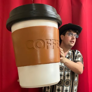 May include: A person in a cowboy hat and glasses holds a giant coffee cup with a brown sleeve that says "COFFEE" in raised letters.