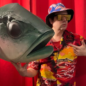May include: A large, green fish head prop with a wide open mouth. The fish head is held in front of a person wearing a Hawaiian shirt, a bucket hat, and yellow-tinted sunglasses. The background is a solid red.