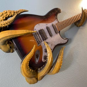 Könnte beinhalten: Eine braune und schwarze E-Gitarre mit einem weißen Schlagbrett ist an einer Wand mit einer gelben, tentakelartigen Skulptur befestigt. Die Gitarre hat einen hölzernen Hals und einen Satz von sechs Stimmwirbeln.