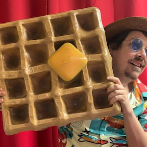 May include: A person wearing a colorful shirt and a brown hat holds a large, brown waffle with a yellow square of butter in the center.