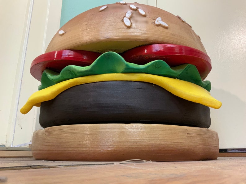 Puede incluir: Una gran escultura de hamburguesa de madera pintada a mano con un pan marr&oacute;n, un tomate rojo, lechuga verde, queso amarillo y una hamburguesa negra.