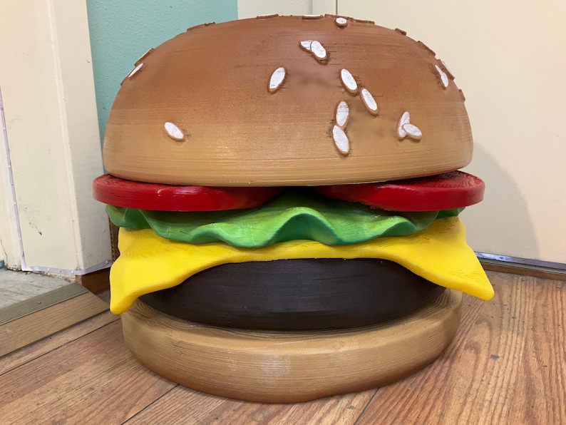 Puede incluir: Una gran escultura de hamburguesa de espuma, de color marr&oacute;n y amarillo, con semillas de s&eacute;samo en el pan superior. La escultura es una representaci&oacute;n realista de una hamburguesa.