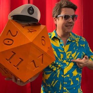 Pu&ograve; includere: Un dado arancione sovradimensionato a venti facce con numeri bianchi, un cappello da marinaio bianco con bordo nero e un'emblema dorato. Il dado &egrave; tenuto da una persona che indossa una camicia floreale blu e gialla e occhiali da sole.