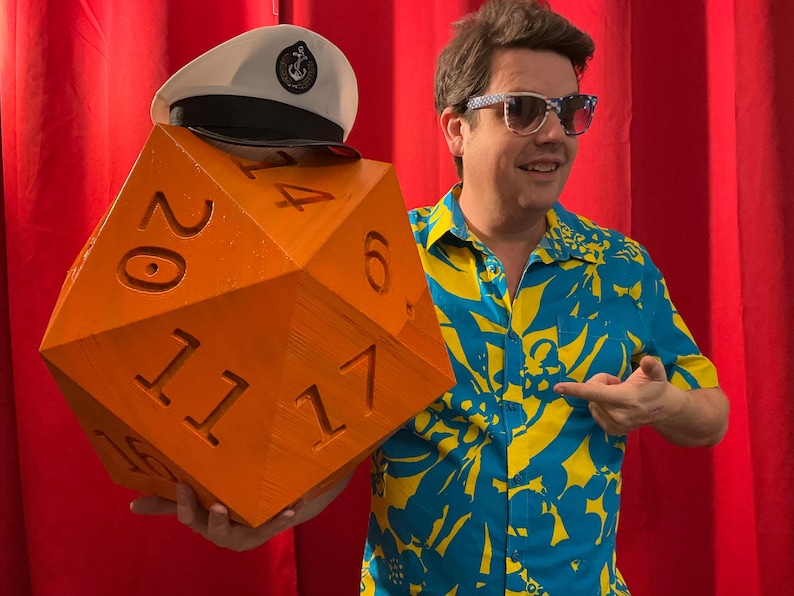 Pu&ograve; includere: Un dado arancione sovradimensionato a venti facce con numeri bianchi, un cappello da marinaio bianco con bordo nero e un'emblema dorato. Il dado &egrave; tenuto da una persona che indossa una camicia floreale blu e gialla e occhiali da sole.
