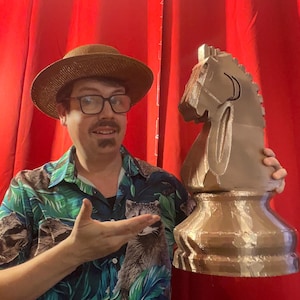 May include: A large, gold-coloured chess knight piece. The knight is held by a person wearing a straw hat, glasses, and a tropical print shirt. The background is a red curtain.