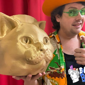 May include: A person wearing a green and yellow Hawaiian shirt and an orange hat with green sunglasses is holding a gold-colored cat head sculpture. The sculpture has a textured surface and large, round eyes. The person is giving a thumbs up and the shirt has the text "Bust Bins" on it.