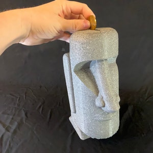 May include: A gray stone-like piggy bank shaped like a human face with a slot on top. A hand is placing a gold coin into the slot.