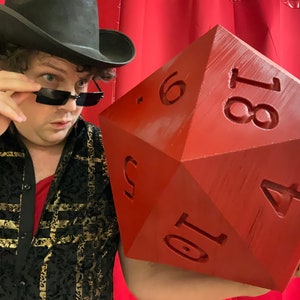 Puede incluir: Una persona con un sombrero negro y gafas de sol sostiene un dado grande, rojo, de veinte caras. El dado tiene los n&uacute;meros 1, 4, 6, 8, 10 y 18 impresos en sus caras.