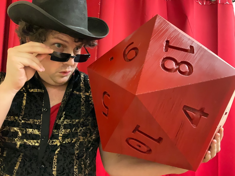 Puede incluir: Una persona con un sombrero negro y gafas de sol sostiene un dado grande, rojo, de veinte caras. El dado tiene los n&uacute;meros 1, 4, 6, 8, 10 y 18 impresos en sus caras.