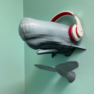 May include: A gray 3D printed whale head with a white and red headphone on top. The whale is mounted on a wall with a gray 3D printed tail fin.