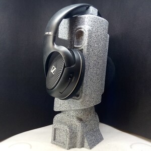 以下が含まれることがあります： 黒色のオーバーイヤーヘッドホン。ヘッドホンは、黒色のテクスチャ加工が施されたイヤーカップと、黒色のヘッドバンドを備えています。ヘッドホンは、灰色のフォーム製のスタンドの上に置かれています。