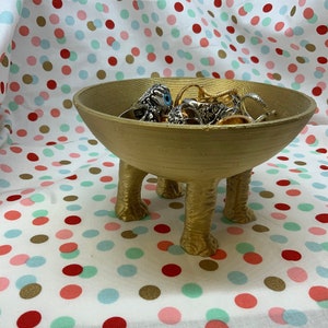 May include: A gold-coloured, three-legged bowl filled with jewellery. The bowl has a rounded shape and is elevated by three short, textured legs. The jewellery includes rings and necklaces. The background is a white fabric with colourful polka dots.