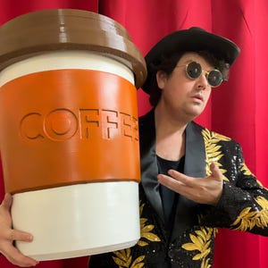 Pode incluir: Uma pessoa vestindo um blazer preto com detalhes em folha de ouro e óculos de sol está segurando uma grande xícara de café branca com uma tampa marrom e uma manga de papel marrom que diz "COFFEE" em letras em relevo.