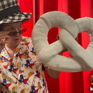 May include: A person wearing a Texas-themed shirt and a checkered hat holds a large, white, soft pretzel. The pretzel has a detailed, textured surface. The background is a red curtain, and the Big Food logo is in the bottom right.
