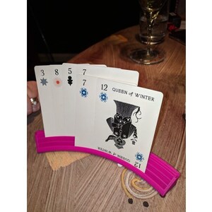 May include: A pink card holder with a curved design holds a set of playing cards. The cards feature a black and white illustration of a queen with a crown and a snowflake design. The cards also have numbers and symbols on them.