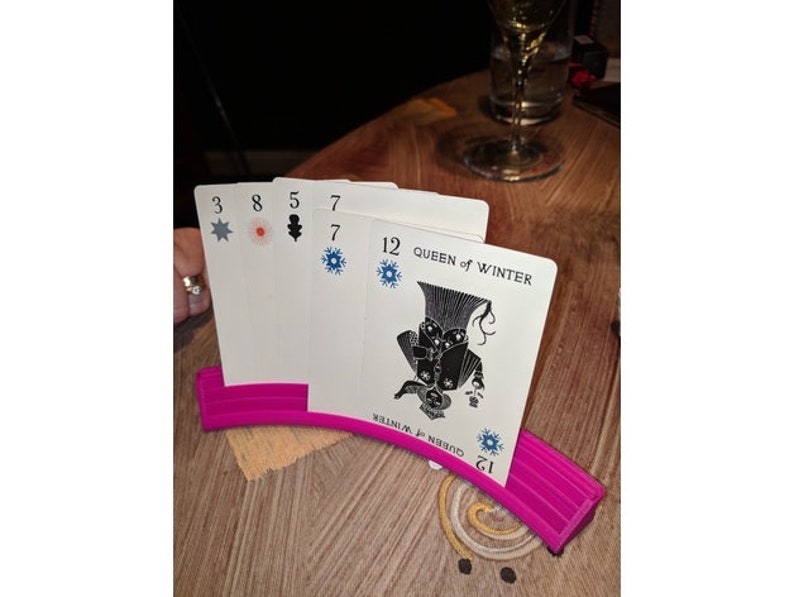 May include: A pink card holder with a curved design holds a set of playing cards. The cards feature a black and white illustration of a queen with a crown and a snowflake design. The cards also have numbers and symbols on them.