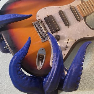Può includere: Una chitarra elettrica sunburst con un battipenna bianco e artigli blu testurizzati stampati in 3D su entrambi i lati.