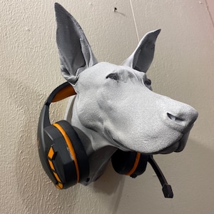 May include: A gray dog head wall mount with large ears, wearing a black and orange gaming headset. The dog's head is textured and mounted on a beige wall, creating a unique display for headphones or a gaming setup.