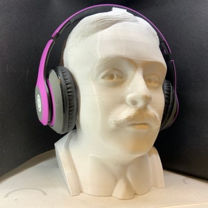 May include: A white 3D printed bust of a person wearing purple and black headphones. The bust has a mustache and is looking straight ahead.