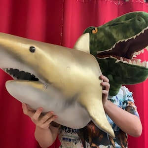 May include: A person wearing a colorful shirt holds a large, gold-painted shark head and a green plush dinosaur head. The dinosaur head is open-mouthed and has sharp teeth.
