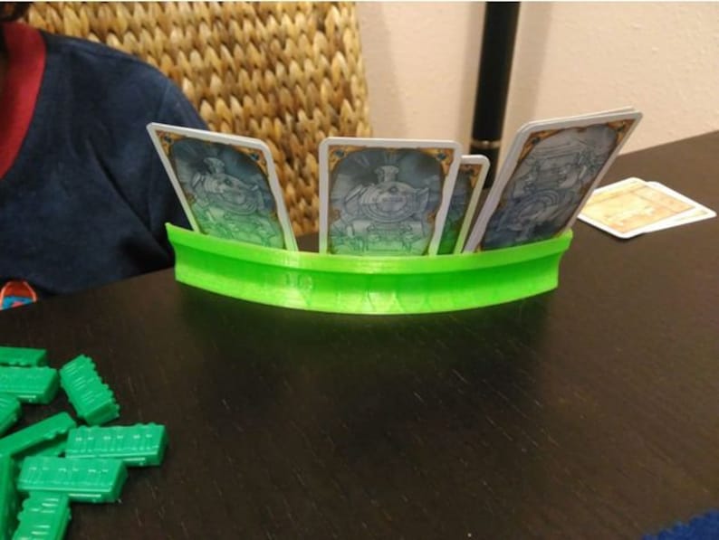 May include: A green card holder with four cards inside. The cards are white with blue and gold designs. The holder is curved and has a smooth surface.