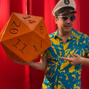 May include: An oversized orange twenty-sided die with black numbers. The die is held by a person wearing a captain's hat, sunglasses, and a blue and yellow patterned shirt. The background is a red curtain.