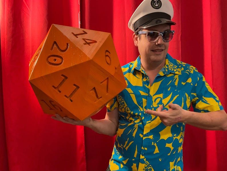 Pu&ograve; includere: Un dado arancione a venti facce di grandi dimensioni con numeri neri. Il dado &egrave; tenuto da una persona che indossa un cappello da capitano, occhiali da sole e una camicia fantasia blu e gialla. Lo sfondo &egrave; una tenda rossa.