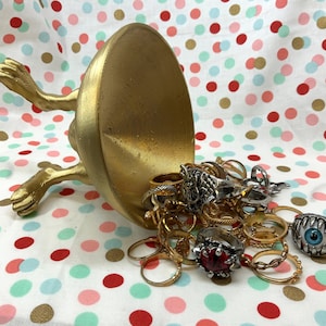 Könnte beinhalten: Ein goldfarbener Ringhalter in Form eines Fusses und einer Schale, überquellend mit verschiedenen Ringen. Die Ringe sind silber- und goldfarben, einige mit Edelsteinen und einzigartigen Designs, vor einem gepunkteten Hintergrund.