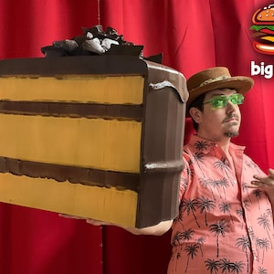 May include: A person wearing a pink shirt with palm trees printed on it and a brown hat with a green brim is holding a large chocolate cake with yellow frosting and chocolate shavings on top. The text "big food" is written in white letters on a black background.