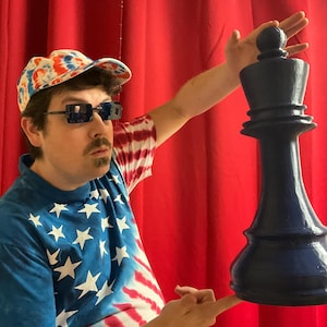Op de afbeelding: Een persoon draagt een rood, wit en blauw tie-dye shirt en een rood, wit en blauw tie-dye pet en houdt een groot, blauw schaakkoningstuk omhoog. De persoon staat voor een rood gordijn.