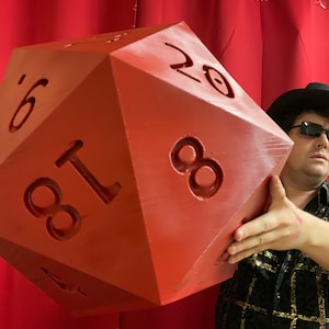 Puede incluir: Un dado grande de veinte caras de color rojo con los n&uacute;meros 1, 4, 6, 8, 11, 18 y 20 visibles. Una persona sostiene el dado frente a una cortina roja.