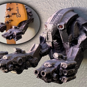 Peut inclure: Support mural pour guitare en métal gris, en forme de robot futuriste. Le robot a de multiples appendices en forme de canon et des détails d'yeux rouges. La tête de la guitare est visible en arrière-plan, tenue par les bras du robot.