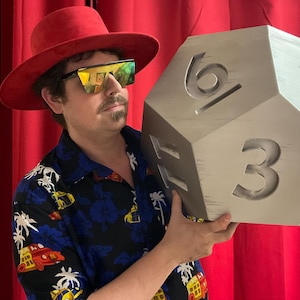 May include: A large, silver-colored, oversized die with the numbers 3, 4, and 6 visible. The die is held by a person wearing a red hat, sunglasses, and a blue Hawaiian shirt with a car and palm tree pattern.