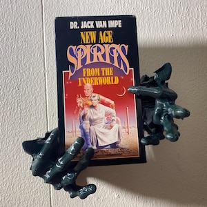 May include: A book titled "New Age Spirits From The Underworld" by Dr. Jack Van Impe, held by a dark green, skeletal hand-shaped book holder. The book cover features a couple in a desert setting.