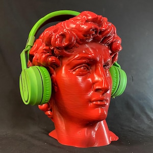 May include: A red bust of David wearing bright green headphones. The sculpture is a vibrant red, with the headphones contrasting in a vivid green. The headphones appear to be over-ear style, adding a modern touch to the classic art piece.