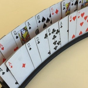 May include: A black plastic card holder with a curved design holds a deck of playing cards. The cards are fanned out in a semi-circle, showing the face of each card. The cards include Ace, Queen, Jack, 10, 5, 4, 2, and Joker.