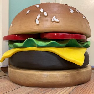 Puede incluir: Una gran escultura de hamburguesa de madera marr&oacute;n con semillas de s&eacute;samo en el pan superior, un tomate rojo, lechuga verde, una rebanada de queso amarillo y una hamburguesa marr&oacute;n.