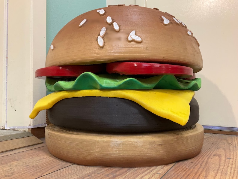 Puede incluir: Una gran escultura de hamburguesa de madera marr&oacute;n con semillas de s&eacute;samo en el pan superior, un tomate rojo, lechuga verde, una rebanada de queso amarillo y una hamburguesa marr&oacute;n.