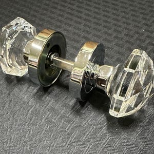 May include: A pair of crystal door knobs with a faceted design. The knobs are clear and mounted on a shiny, silver-colored metal base. The door hardware is designed for interior doors.